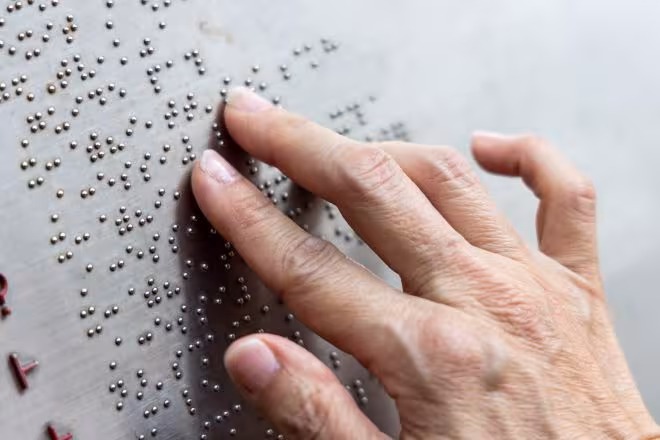 Braille írás látszik a képen. Közelről, egy kéz és az újak éppen letapogatják az írást. 